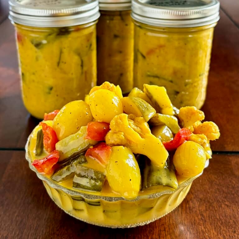 Newfoundland Mustard Pickles photo of mixed pickles in an antique clear glass serving dish with bottles of finished pickles in the background. Photo is cropped square as a Google Featured Image.