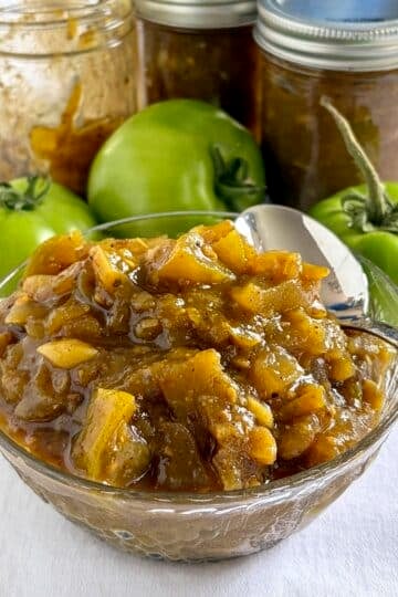 Close up photo of Photo of green tomato chow chow in a glass serving bowl with green tomatoes in the background for Google Featured Image.