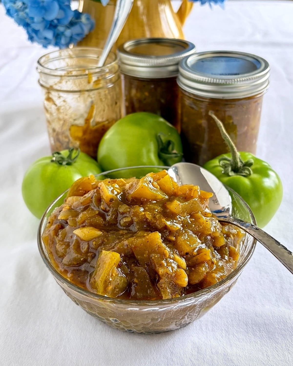 Photo of green tomato chow chow in a glass serving bowl with green tomatoes in the background.