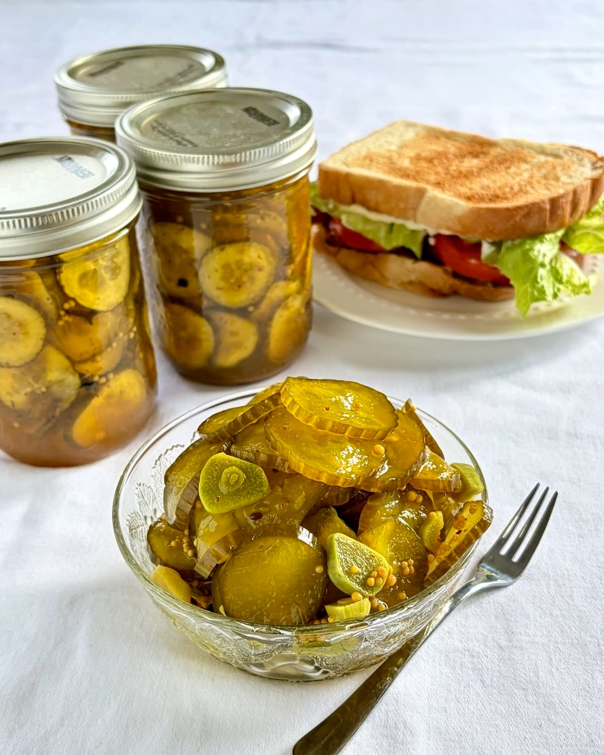 Photo of Sweet Yum Yum Pickles in a serving bowl with pickle bottles and sandwich in the background.