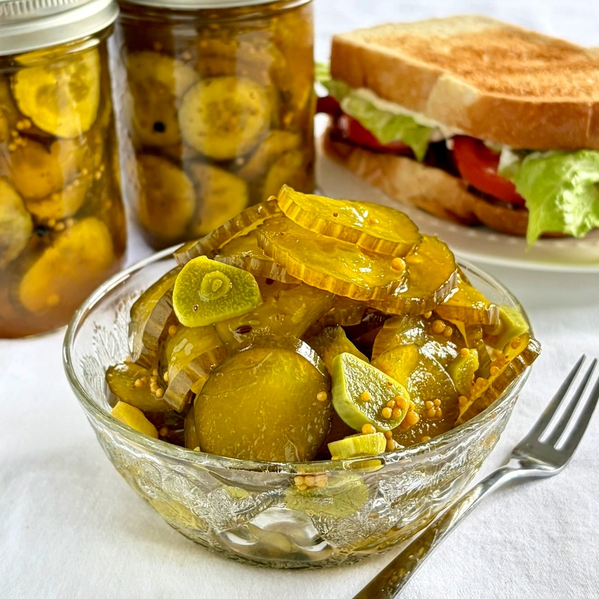 Close up photo of Photo of Sweet Yum Yum Pickles in a serving bowl with pickle bottles and sandwich in the background.