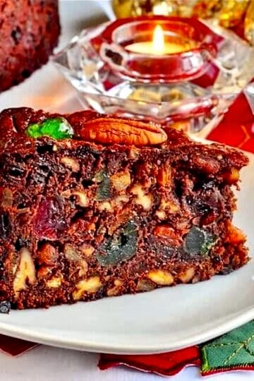 Close up photo of one slice of Old English Fruitcake on a white plate with Christmas decorations in background.