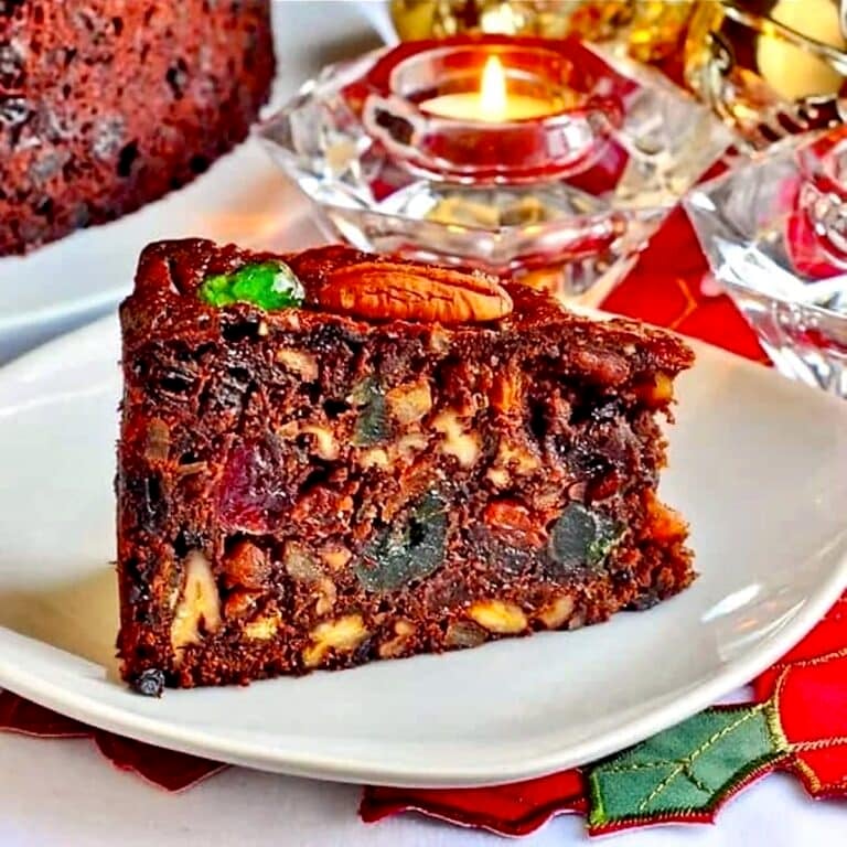 Close up photo of one slice of Old English Fruitcake on a white plate with Christmas decorations in background.